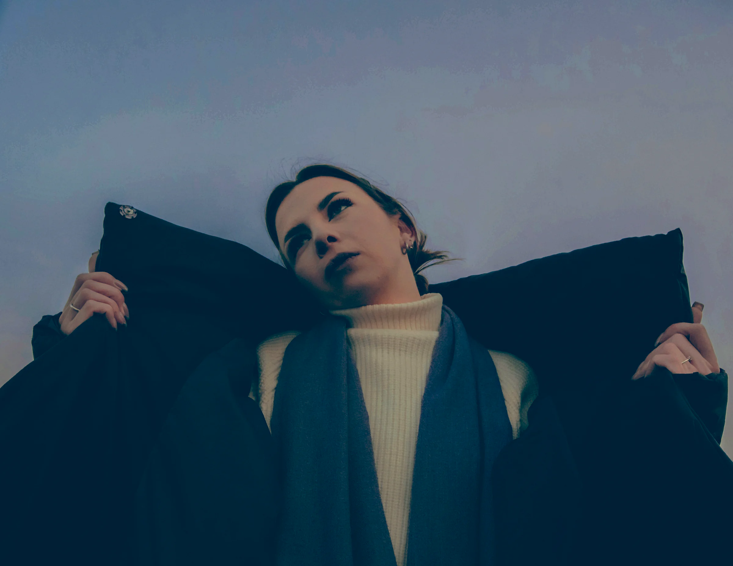 A woman holding a black coat up around her shoulders against a pale grey sky.