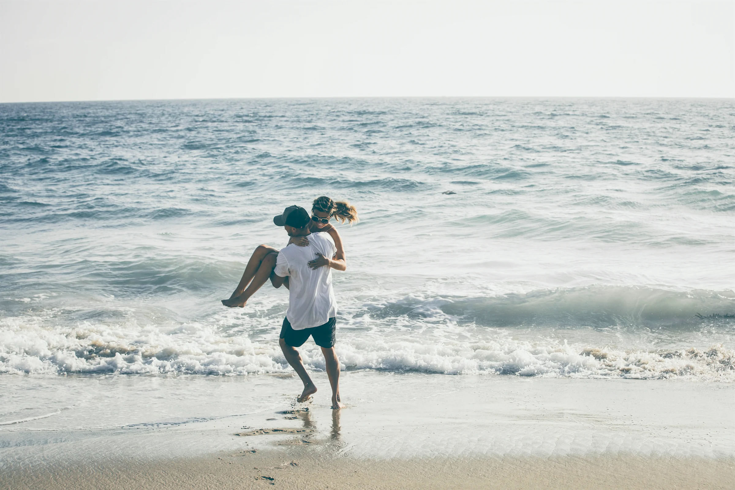 A man carrying a woman through the surf on a bright, hazy beach.